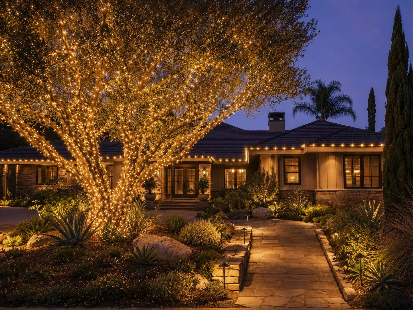 Mature deciduous tree wrapped in warm-white Christmas lights at the base of the trunk, single-story ranch home behind