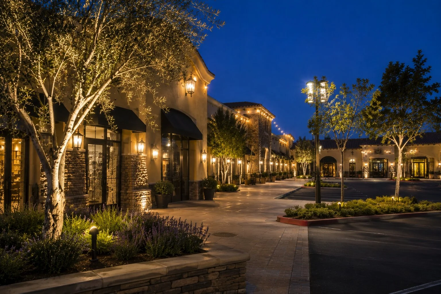 Spanish revival retail main street at dusk with downlight lanterns on storefronts, olive uplighting between, and street lamps