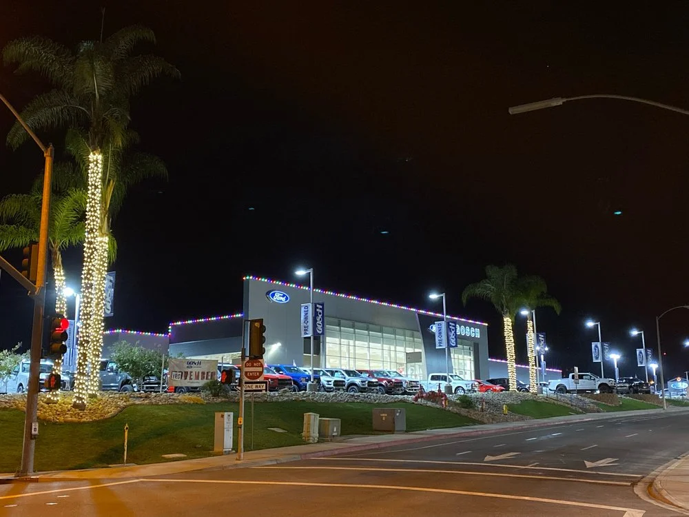 Ford dealership at night with multicolor Christmas C9 lights along the roofline and warm-white wraps on the front palm trees