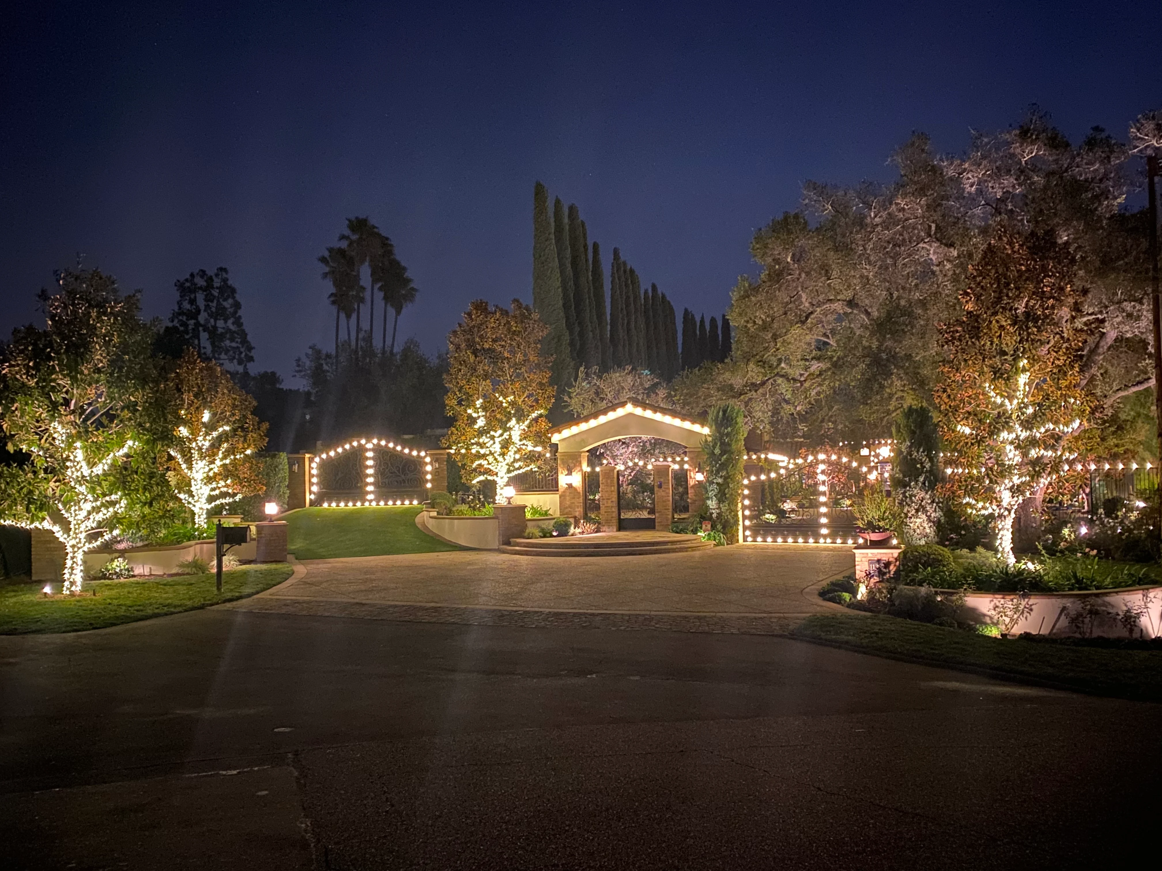 Estate driveway entry with warm holiday lighting across trees and gate
