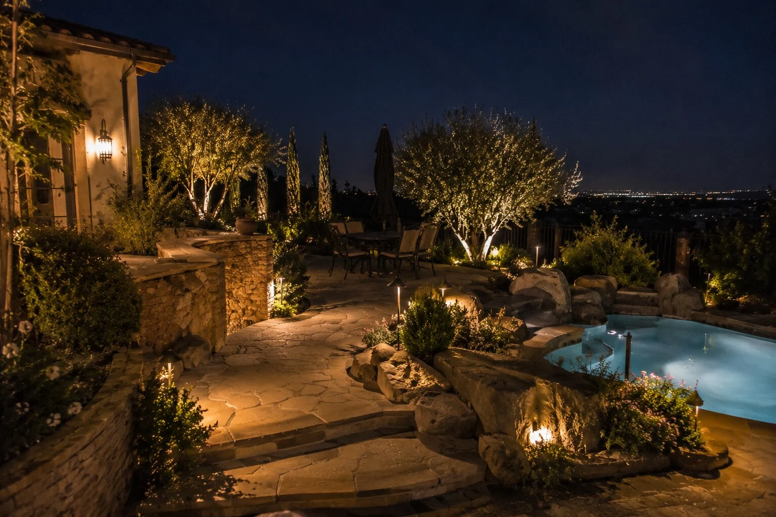 Backyard pool view at dusk with architectural uplighting on the residence, palm and olive uplights along the perimeter