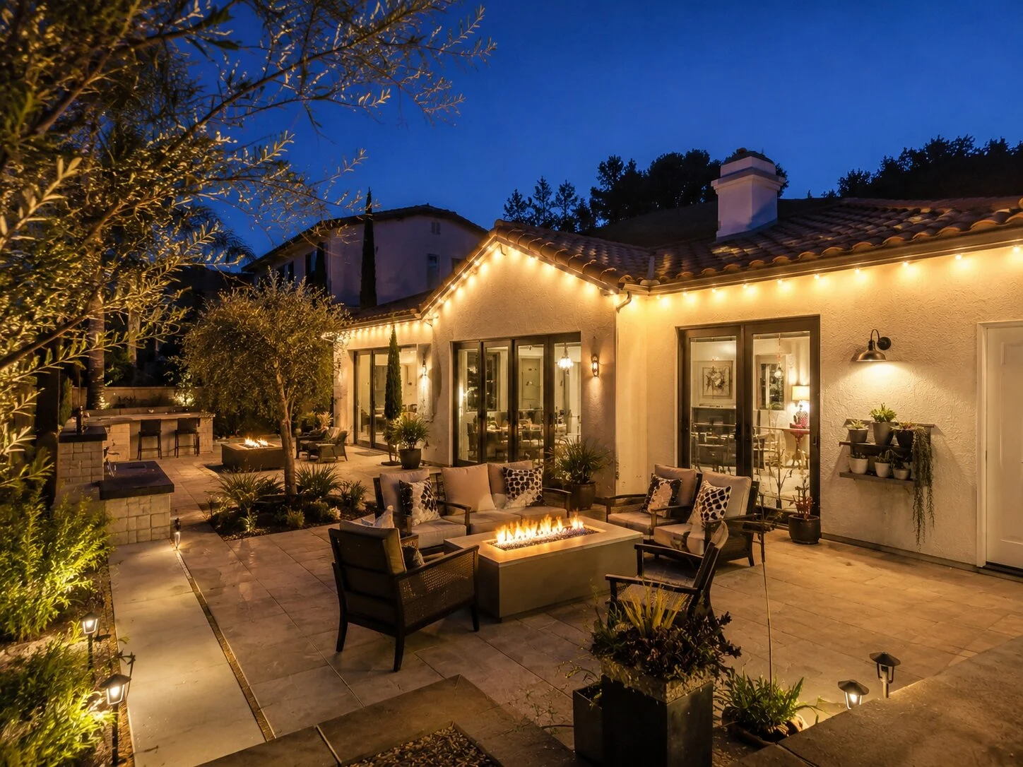 Spanish home with patio firepit and lounge at dusk, warm-white permanent eave lighting on every roof line, olive trees flanking the patio