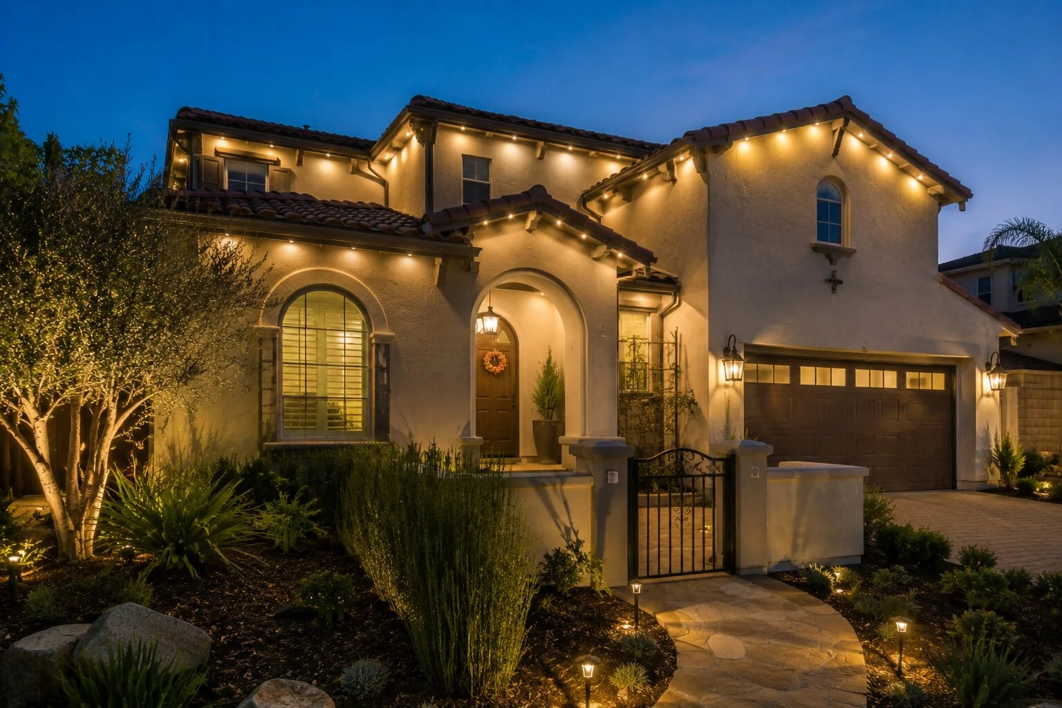 Two-story Spanish home with arched windows and path-lit garden beds at dusk, warm-white permanent eave lighting along every gable