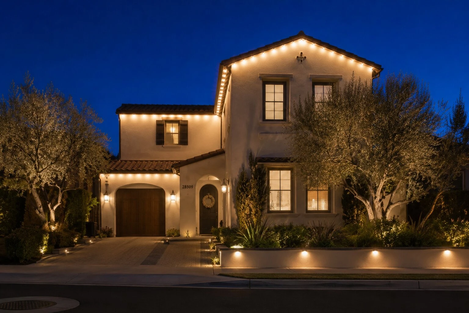 Two-story symmetric Spanish revival home at dusk with warm-white permanent LED lights running under every eave and gable, two olive trees flanking the entry, paver driveway and path lights in the planter beds