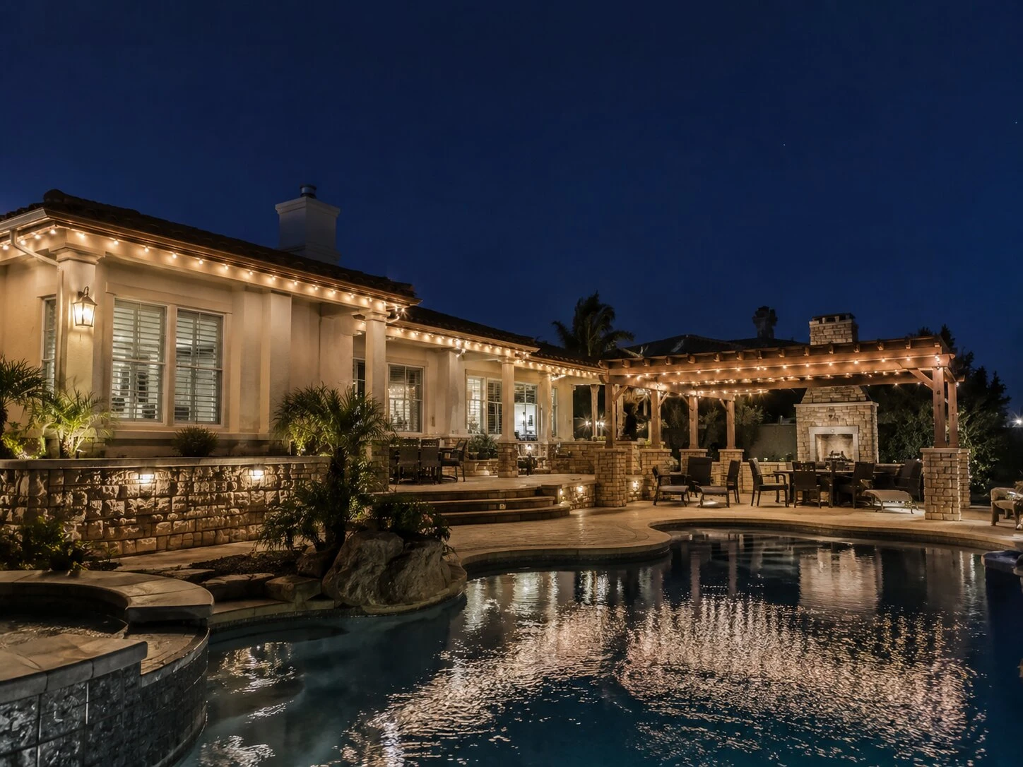 Large Spanish home at deep-blue dusk with a covered pergola and stone fireplace at right, warm-white permanent LED lights along the eave and pergola, swimming pool with the lights reflected in the foreground
