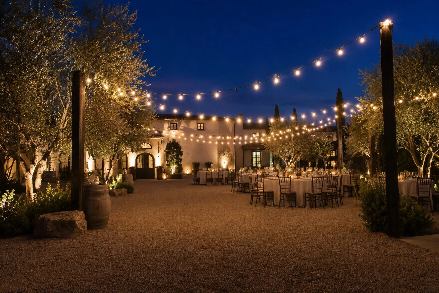 Spanish revival courtyard reception at dusk with round farmhouse tables and chiavari chairs on a gravel patio, bistro string lights overhead between olive trees and an arched stone entry