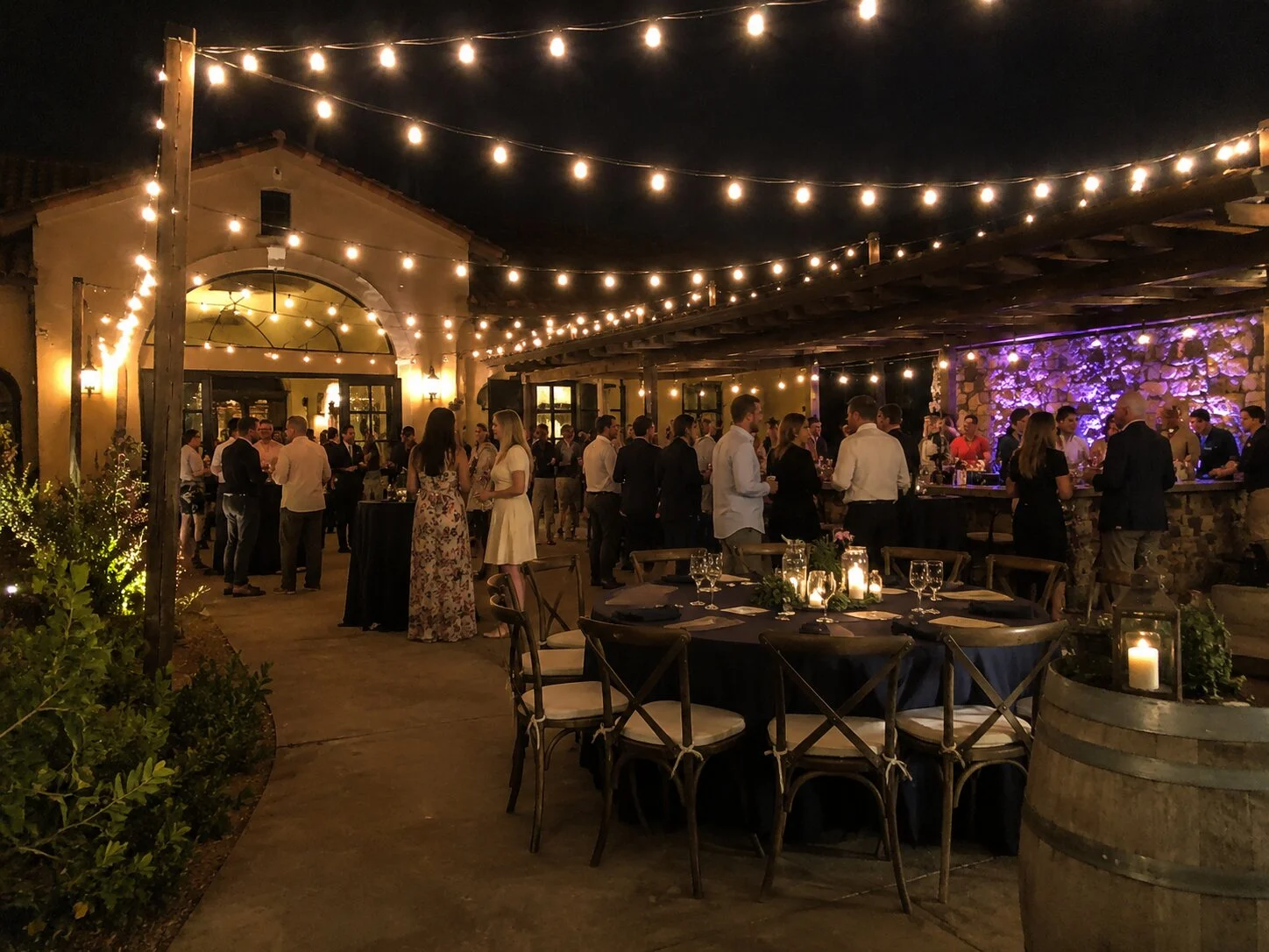 Cocktail reception at night with mingling guests, bistro string lights overhead, Spanish revival archway and a stone bar with purple accent uplighting, dressed cocktail tables