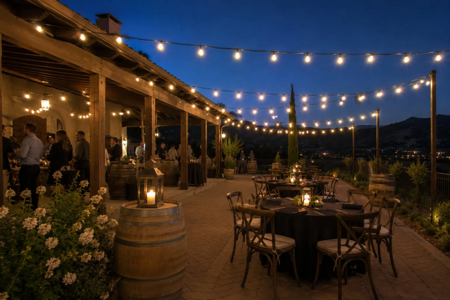 Wine-country terrace cocktail reception at dusk with mingling guests on a paver patio, bistro string lights overhead, wine-barrel cocktail tables with lanterns, mountain valley view